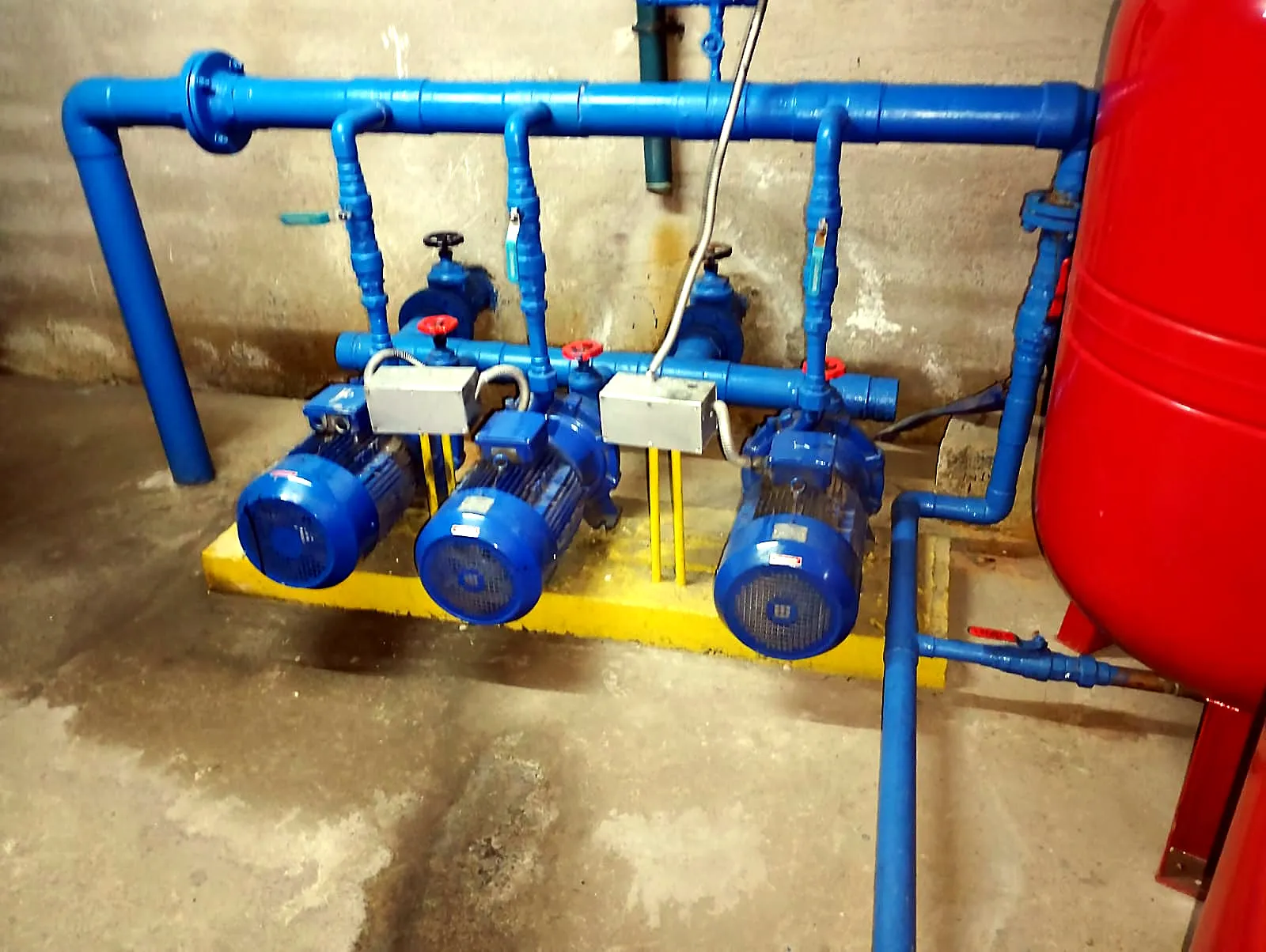 Grupo de tres motobombas azules con su colector y estanque hidroneumático rojo en la sala de bombas de un edificio